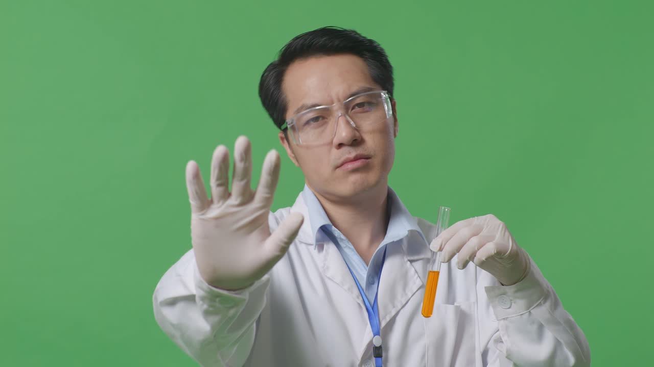 실험관에 오렌지색 액체가 들어있는 아시아 남성 과학자가 카메라를 바라보고 실험실의 녹색 스크린 배경에 손 표시 없이 불쾌감을 표합니다.