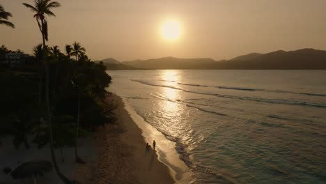 Couple-on-a-paradise-beach-surrounded-by-mountain,-sunset,-aerial-rise-up