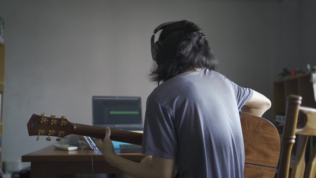 A man with long hair wearing headphones and playing acoustic guitar in order to recording into a computer in a domestic room