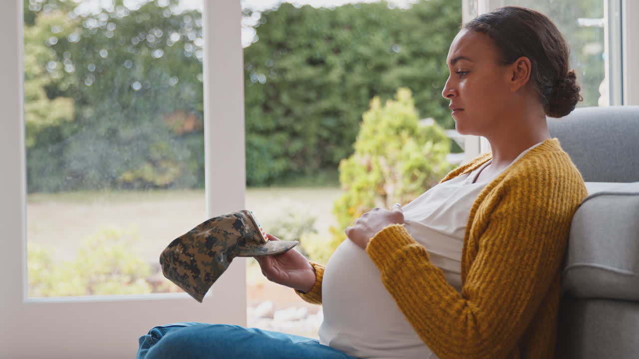 Lonely Pregnant Woman Sitting On Floor At Home Holding Army Husband's Cap