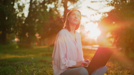 lady in pink top with glasses resting on her head smiles warmly while typing on laptop, she looks up thoughtfully, then refocuses on her laptop, with sunlight creating a beautiful glow around her