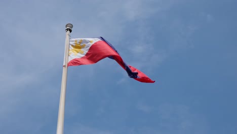 bandera nacional filipina como la cámara se inclina hacia arriba y se aleja un poco revelando la bandera volando hacia la derecha y el fantástico cielo azul y algunas finas nubes esponjosas