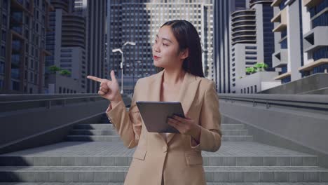an asian business woman using tablet and pointing to the side on the city street