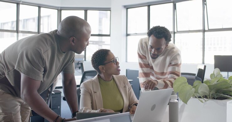 Team leaning in as left coworker showing tablet while woman typing at desk reviewing laptop charts