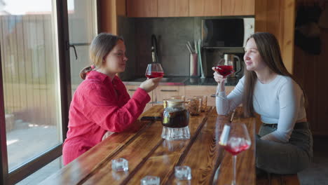 teen girl in red shirt sips from wine glass at wooden table beside another young person indoors near window with soft daylight as beverage pitcher and glasses rest on polished surface