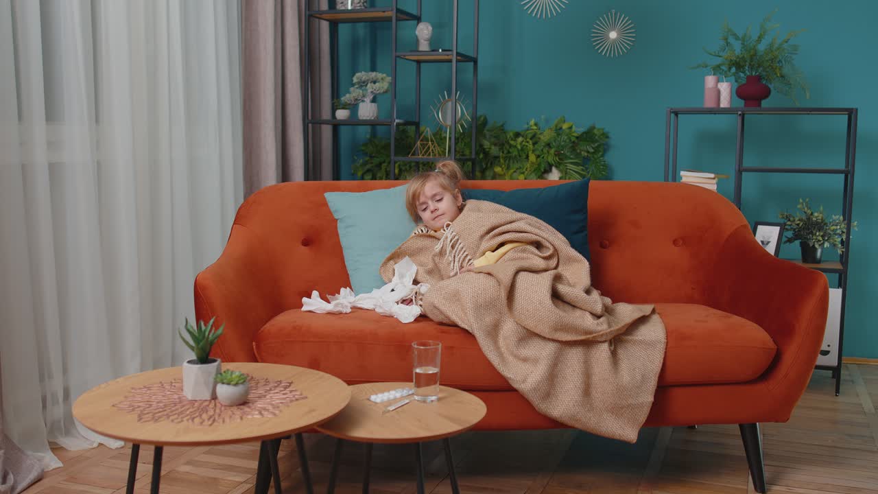 Child girl in plaid suffering from cold fever allergy blows nose snot into napkin on sofa at home