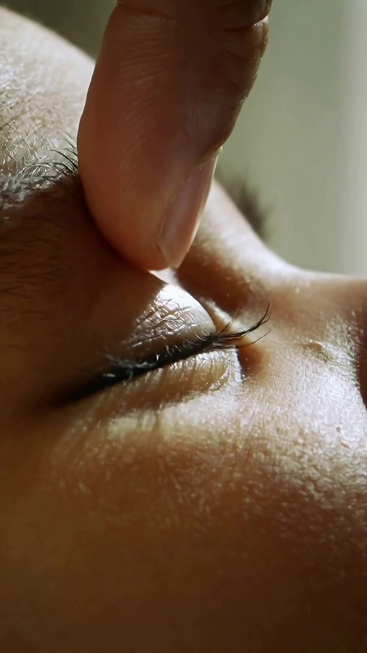 Vídeo vertical: Ponta do dedo se aproximando, pinçando e levantando cílios sobre a pálpebra fechada em luz suave.