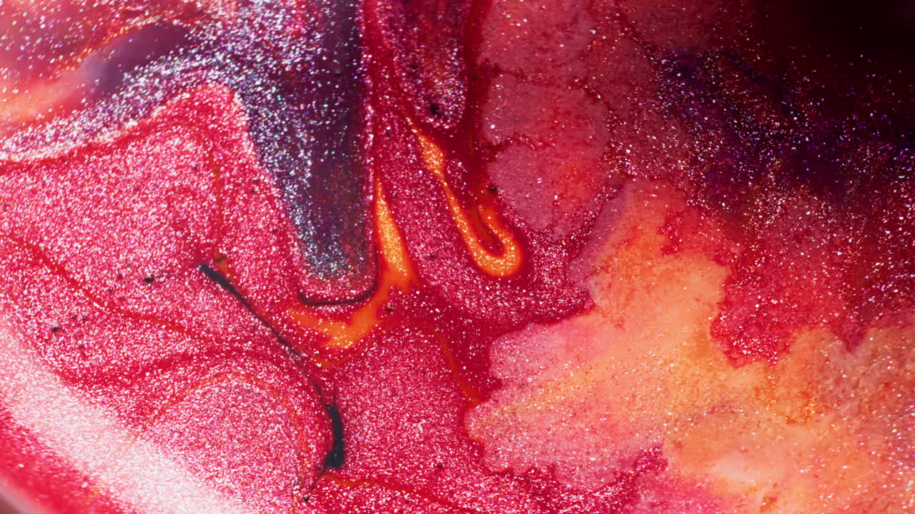 Macro shot of red and orange glitter inks in liquid, as a large drop of purple glitter ink falls and spreads across the surface