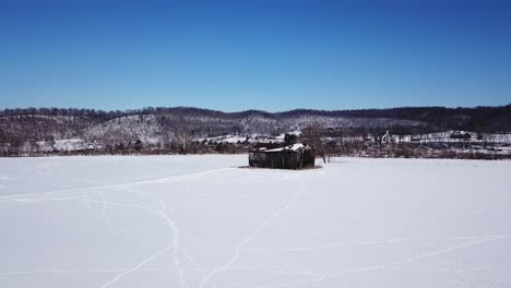 Slow-dolly-aerial-moving-towards-a-large-abandoned,-broken-down,-black-barn-during-the-winter-with-snow-all-over-the-ground-and-animal-tracks-visible