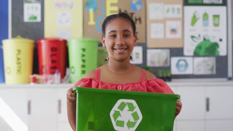 Mixed-race-schoolgirl-smiling,-holding-recycling-bin,-standing-in-classroom