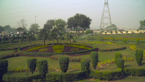 The-impulse-sprinkler-spraying-the-garden,-Beautiful-design-of-the-garden-with-various-plants-and-trees,-Roads-at-the-back-of-the-park,-Traffic-is-going-on