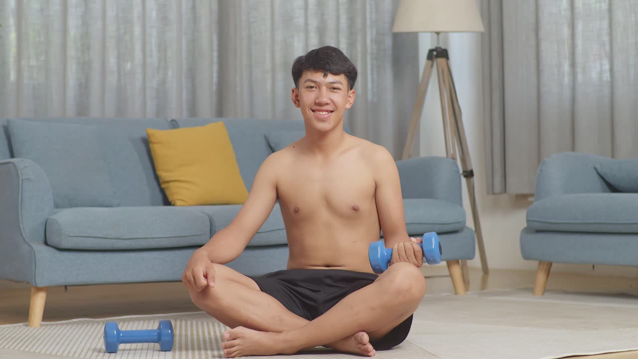 집에서 셔츠를 벗고 운동하는 동안 매트에 앉아 카메라를 향해 미소 짓는 덤벨을 가진 아시아 십대 소년
