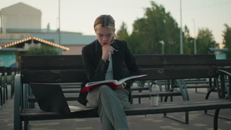 student seated outdoor deeply focused on her open book holding pen near lips with laptop on bench and coffee cup beside her in peaceful urban setting surrounded by trees, benches, and soft lighting