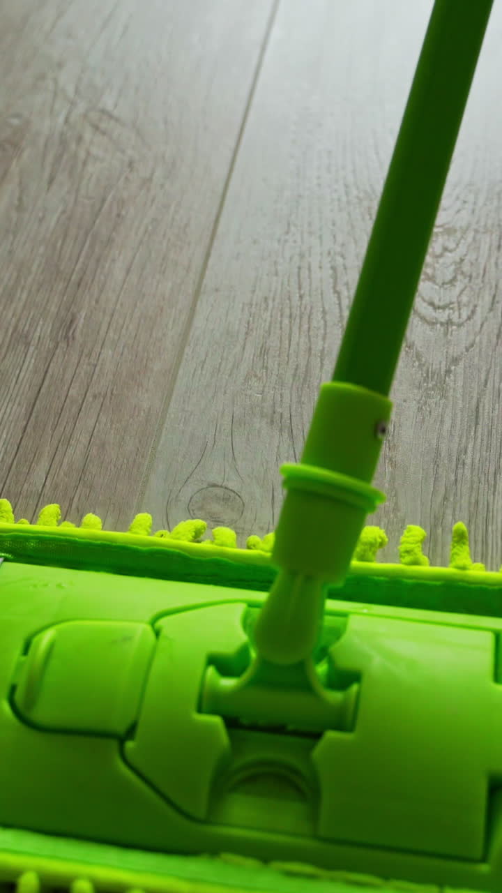 Microfiber wet mop moving around the floor at home. Person in slippers mopping the floor with a green brush. Housework. Close-up. Vertical video