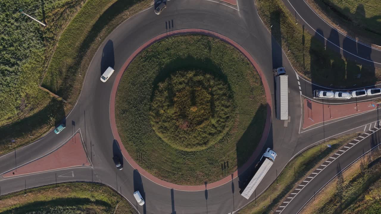 A top-down aerial video of a highway roundabout with trucks and cars moving along the lanes while drone rising to the sky. The circular road is surrounded by green fields and autumn foliage