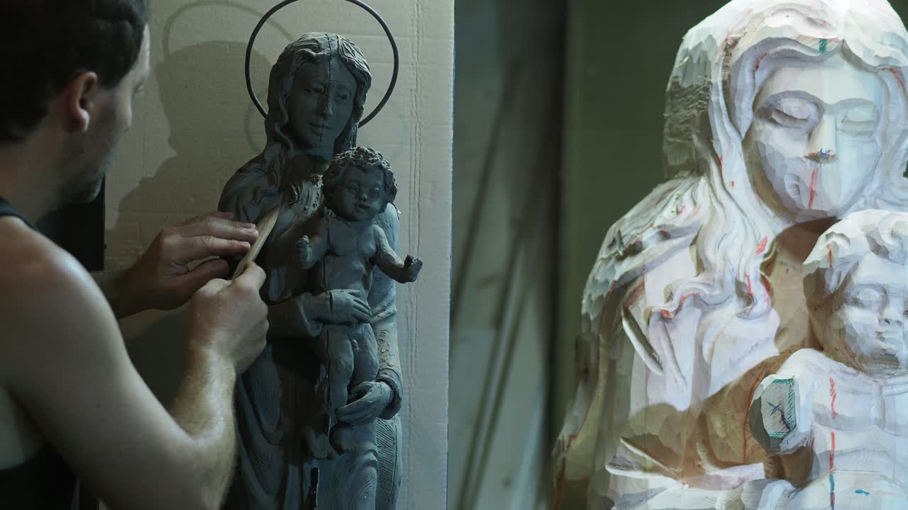 Artist refines small clay model of the Virgin Mary and Child while referencing larger wooden statue near, both sculptures part of a detailed handcrafting process in workshop, slow motion shot