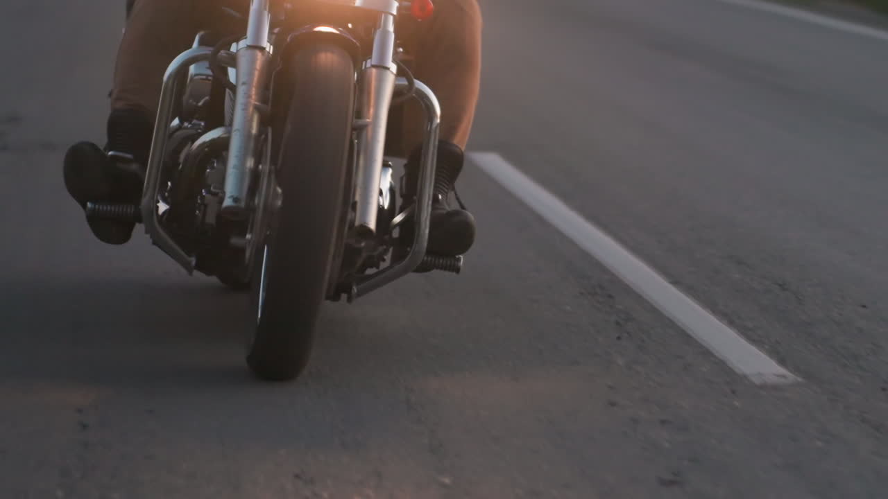 Primo piano dal basso della ruota anteriore di una moto mentre il motociclista si muove su una strada rurale di sera, con il faro che brilla intensamente, movimento che enfatizza velocità, controllo, avventura e libertà di viaggiare all'aperto