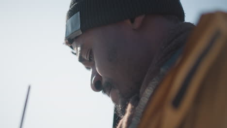 close up aerial view of black man wearing winter coat beanie and gloves sipping coffee through straw while bending head in focus with camera strap visible snowy urban street soft cold winter sunlight