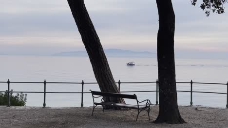 Boat-passing-by-sea-shore-Resort-town-bench-island-in-distance-close-up-Lovran,-Opatija,-Croatia