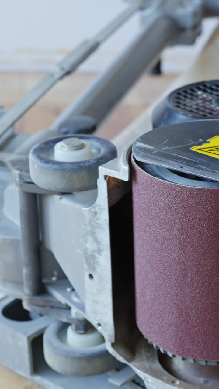 Vertical closeup of industrial floor sanding machine as worker prepares to review and inspect the sandpaper roll, with visible warning label, machine parts, and sanding belt, real time.