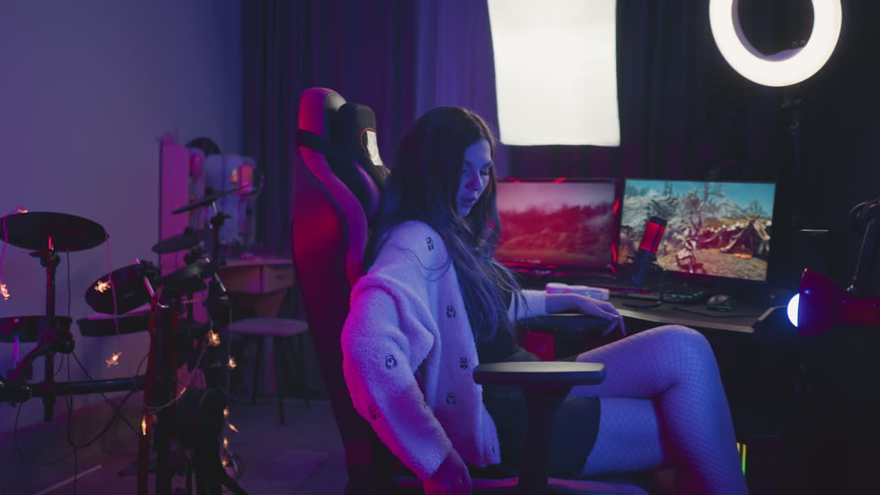 Side view of young woman wearing fishnet tights reclining in gaming chair to relax during gaming session in neon illuminated studio, surrounded by monitors, mic, drum kit, and decorative lighting