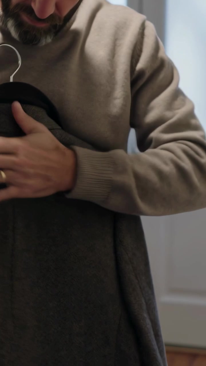 Video verticale: Uomo adulto con maglione beige che solleva un cappotto scuro, controllandone la vestibilità all'ingresso di casa.