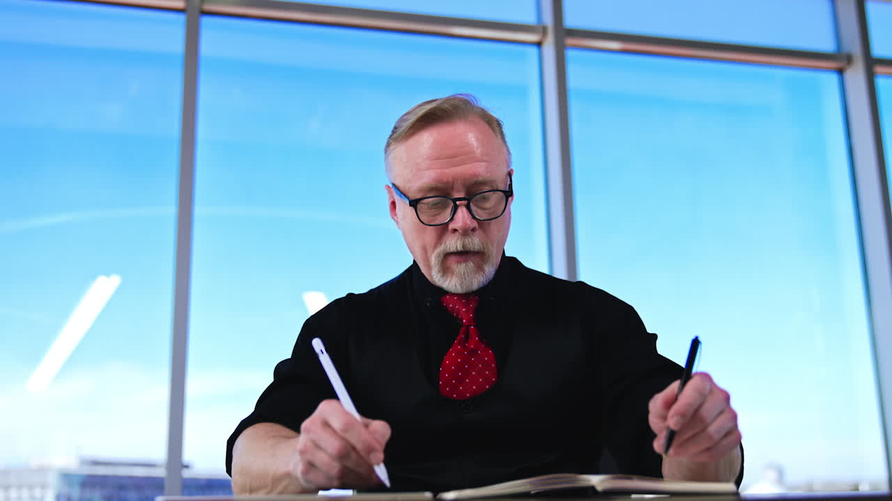 Caucasian man sits at desk holding pens in both hands. Adult man trying to white with both hands. Low angle view.