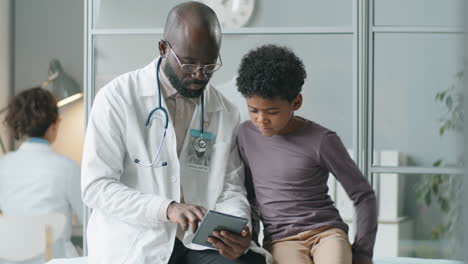 Afroamerikanischer-Arzt-Und-Kleiner-Junge,-Der-In-Der-Klinik-Ein-Tablet-Benutzt