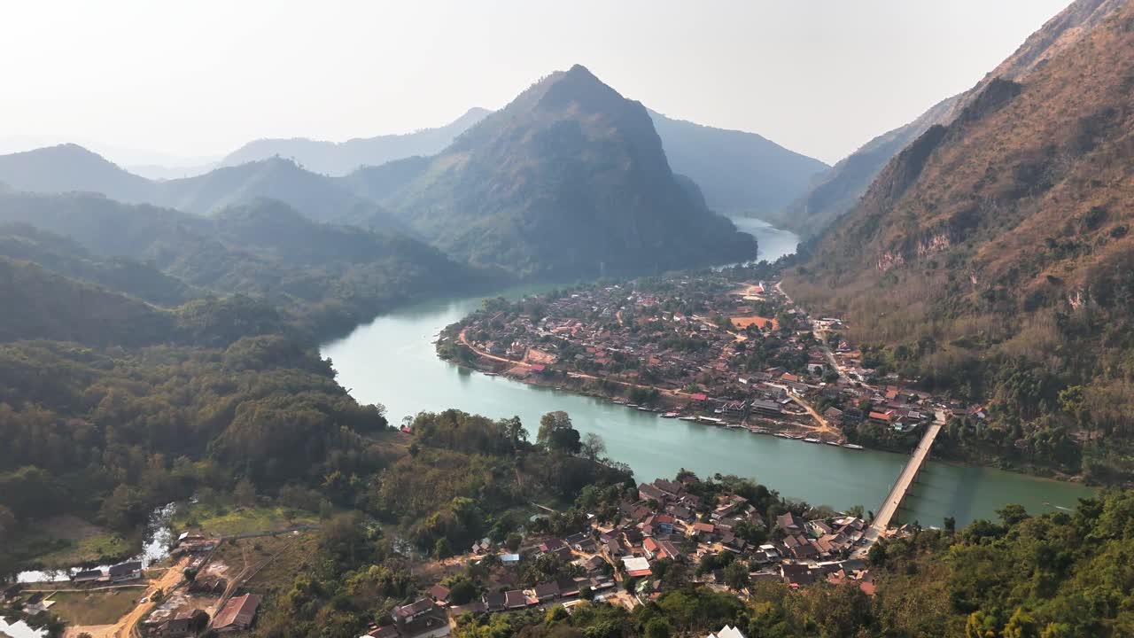 Aerial view of Nong Khiaw, Laos, with a scenic riverbend surrounded by lush mountains, a connected bridge, and traditional village homes nestled in a serene natural valley landscape