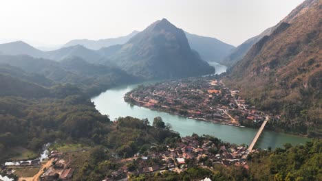 Aerial view of Nong Khiaw, Laos, with a scenic riverbend surrounded by lush mountains, a connected bridge, and traditional village homes nestled in a serene natural valley landscape