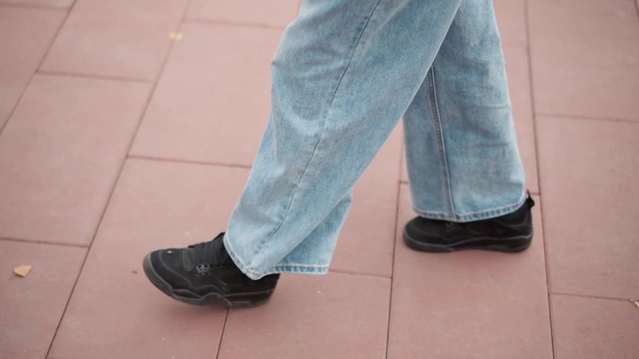 Primo piano delle gambe e dei piedi di una persona che cammina su un pavimento piastrellato rosa con foglie gialle sparse. La persona indossa jeans azzurri a gamba larga e sneakers nere, ripresi dal polpaccio in giù mentre si muove