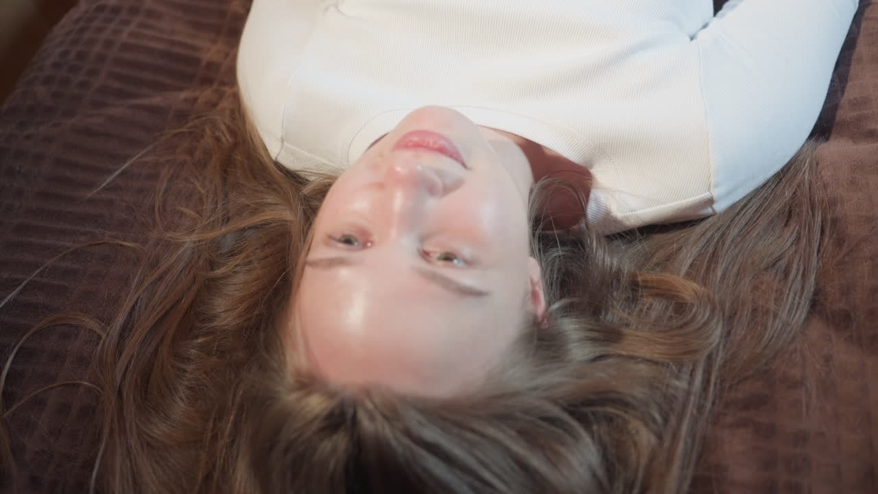 young woman lays on brown bedspread with hair spread out, gazing upward in thoughtful mood, natural light highlighting face and creating soft atmosphere of reflection, peace, and quiet introspection