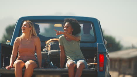 two women riding in back of pick up truck as friends arrive at countryside cabin on road trip