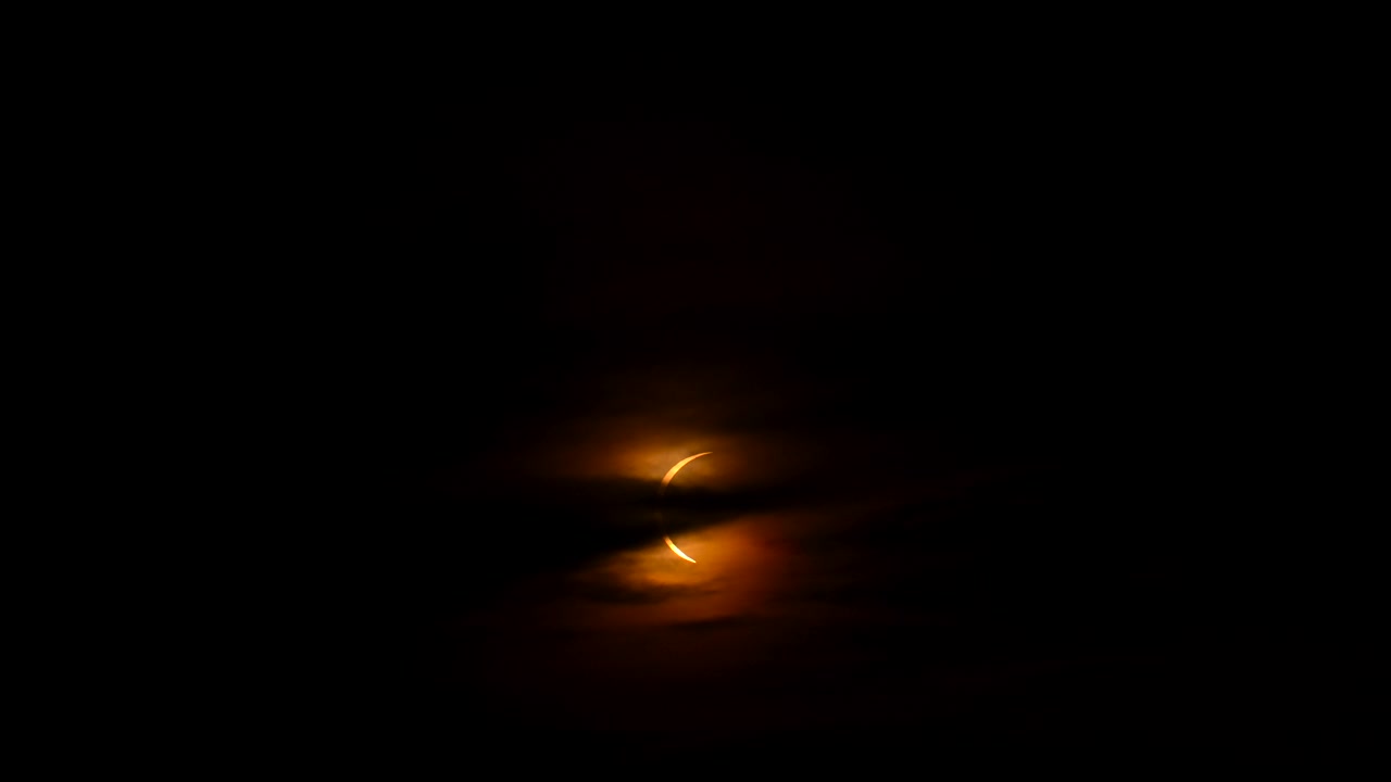 Orange halvmånesol dukker frem bag lagdelte mørke skyer, med detaljer om halvmåne, sol, formørkelse og skyer, der viser himmel, mørke, glød og orange nuancer, med tydelige synlige detaljer, der udgør en kommerciel visualisering.