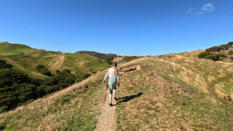 niña caminando por un camino en un día soleado a través de un paisajístico terreno montañoso en nueva zelanda, disparado por un dron siguiendo detrás
