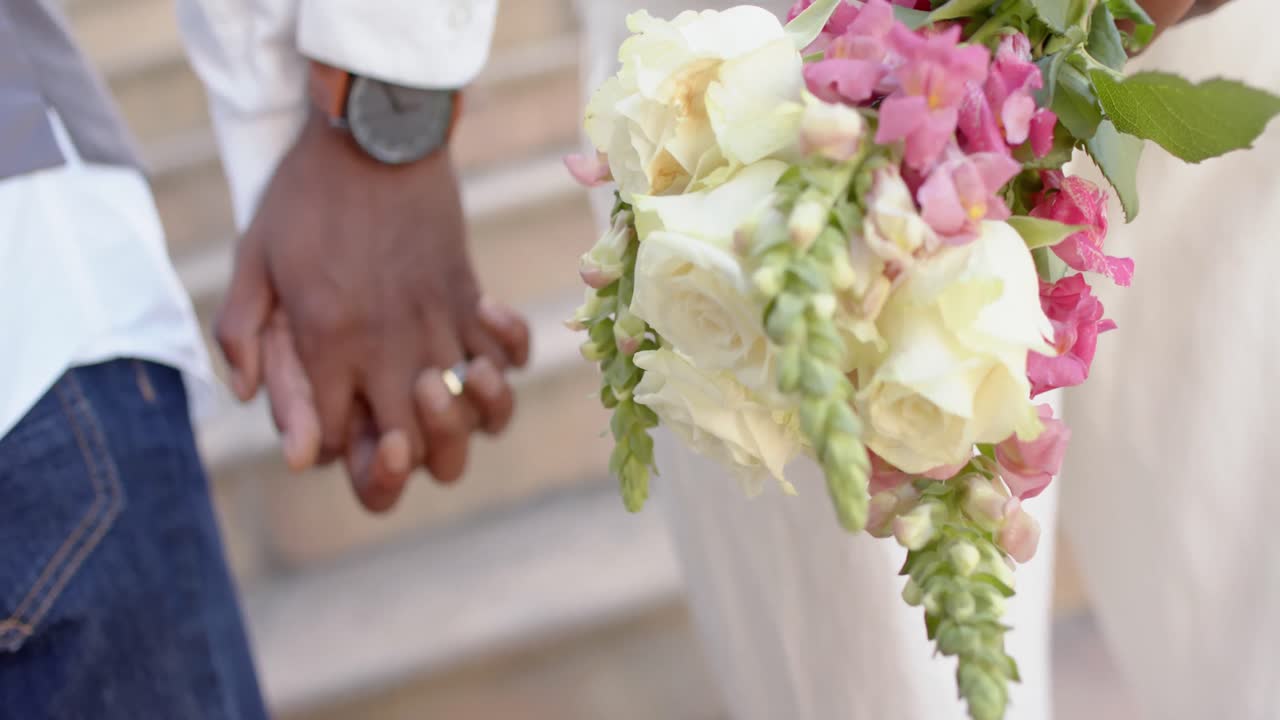 Midsection of african american gay male couple holding hands and flowers at wedding, slow motion