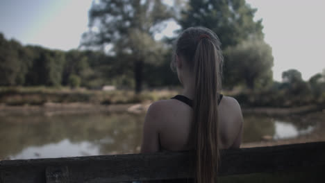 Backside-of-the-young-woman-sitting-under-the-shade-of-the-tree-looking-at-the-still-lake-in-the-distance