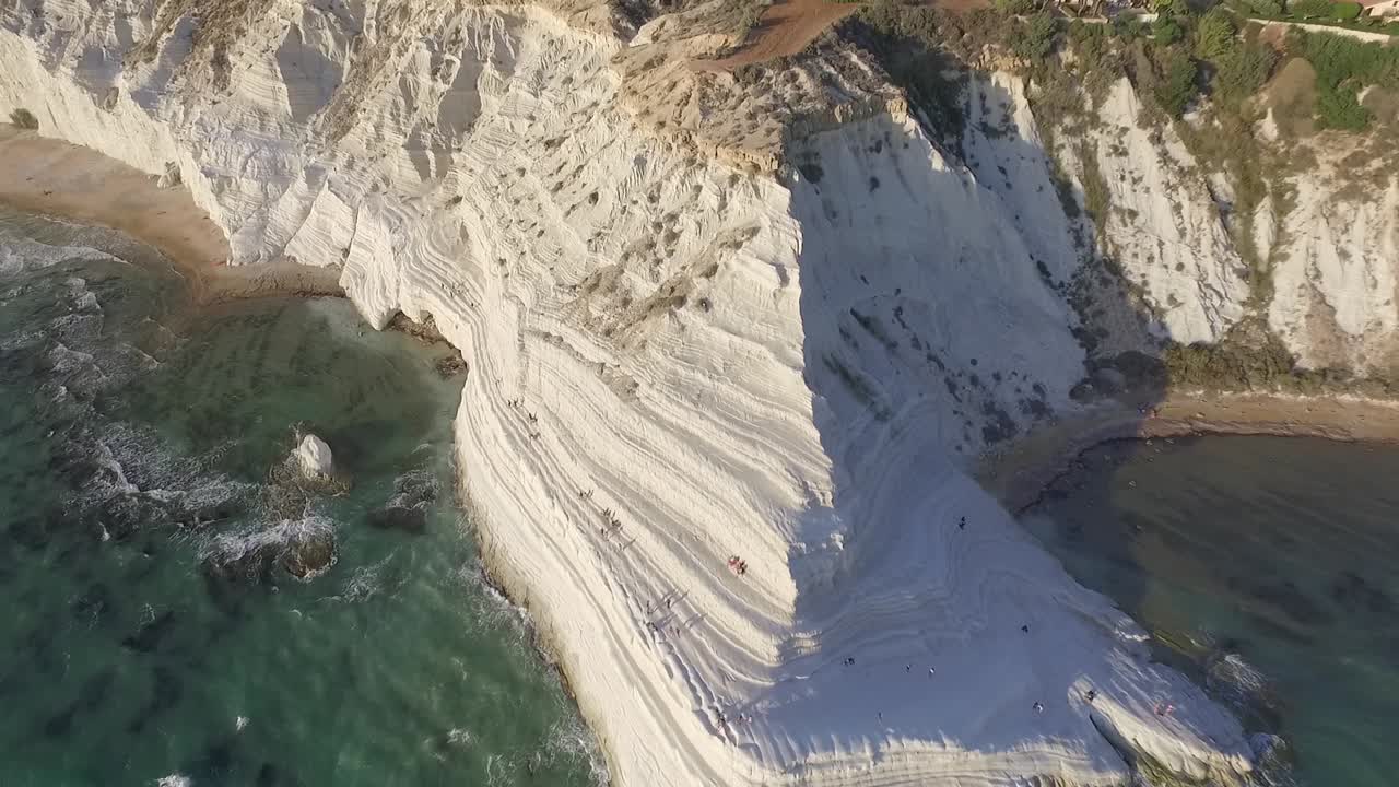 Primo piano aereo delle scogliere di marna bianca della Scala dei Turchi che si tuffano nelle acque turchesi del Mediterraneo, Sicilia, Italia, con formazioni rocciose stratificate drammatiche e onde marine.