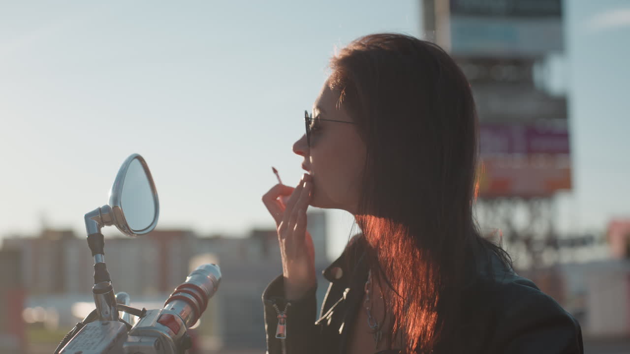 Confident woman in sunglasses and leather jacket applying lipstick while checking motorcycle mirror, with sunlight creating natural glow on face and hair