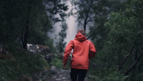 toma en cámara lenta de un hombre con una chaqueta roja corriendo hacia una gran cascada en un bosque