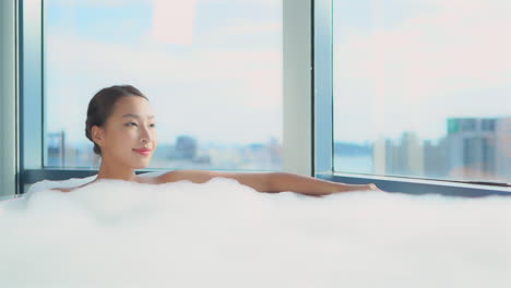 Young-asian-woman-taking-a-hot-bath-with-white-foam-covering-her-body,-admiring-the-city-view-and-relaxing