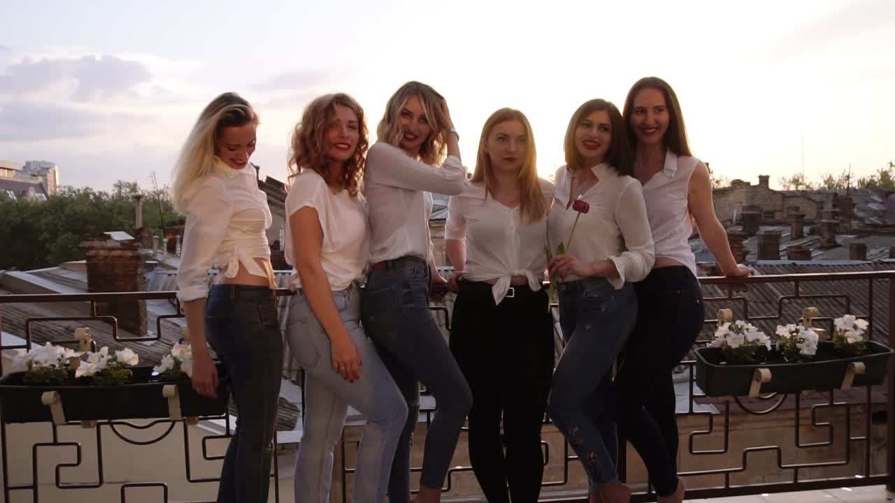 Six caucasian girls are standing on a balcony and posing for the camera. Casual clothes. Close up of girl's legs in jeans. Old city background