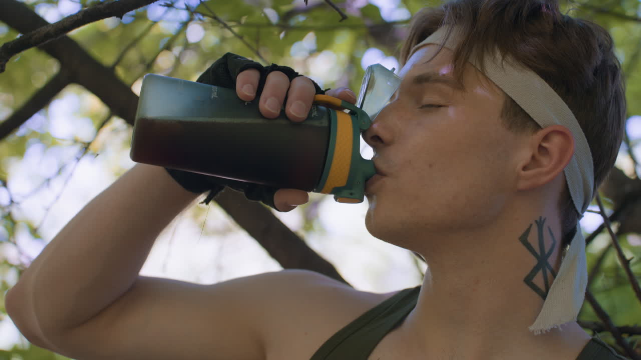 soldier opening eyes and belching in relief while sipping coffee from water bottle under forest canopy with sunlit leaves and branches creating restorative outdoor pause before resuming training