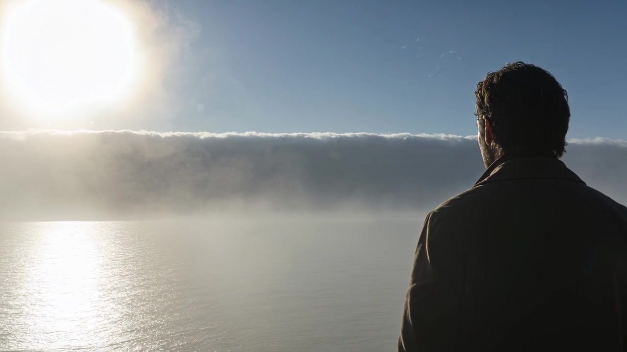 Standing man in dark coat watching low sun dipping over cloud bank and sea, copy space