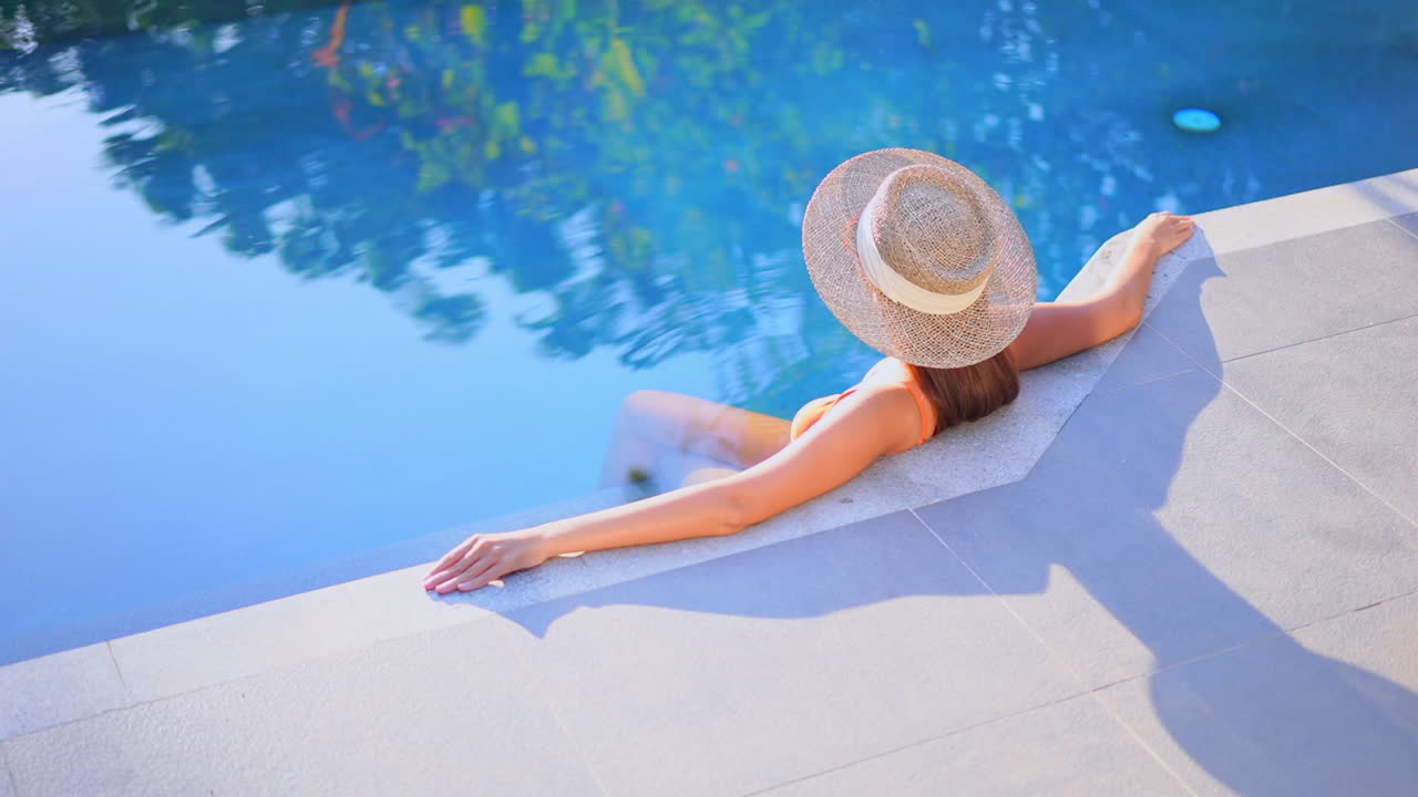 Sexy woman sitting on edge of swimming pool of exotic hotel wearing straw hat