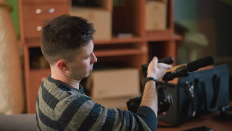 man with back to camera adjusting video equipment in home studio. green wall, soft lighting, and modern furniture in background. focused on setup with microphone and camera gear on table