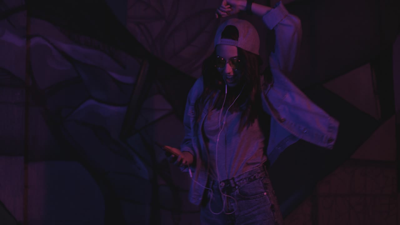 Teenager caucasian girl wearing glasses and cap with headphones while listening to the music and dancing outdoors at night