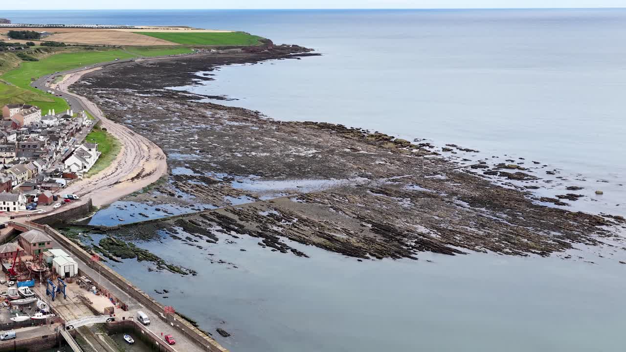 Drone pans above harbor, fishing boats, rocky shoreline, and village under soft daylight in Scotland