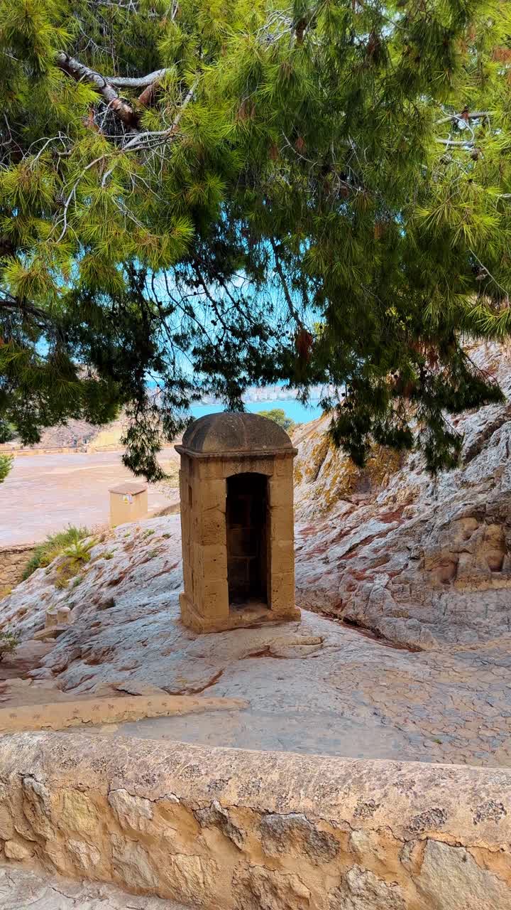 Storica garitta in pietra su scogliera rocciosa a picco sul mare. Tradizionale garitta del Castello di Santa Barbara ad Alicante incorniciata da rami di pino e dal Mar Mediterraneo.