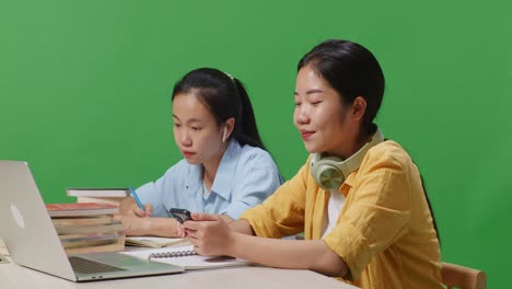 close up of asian woman student wearing headphones and using smartphone while sitting with her friend studying on a table with laptop in the green screen background classroom